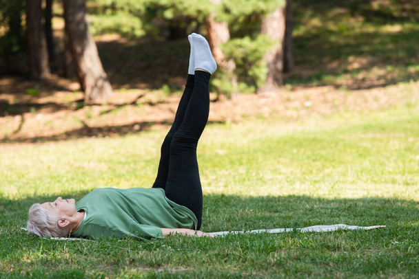 full length of senior woman with grey hair doing shoulder stand on fitness mat in park - Foto, Imagem