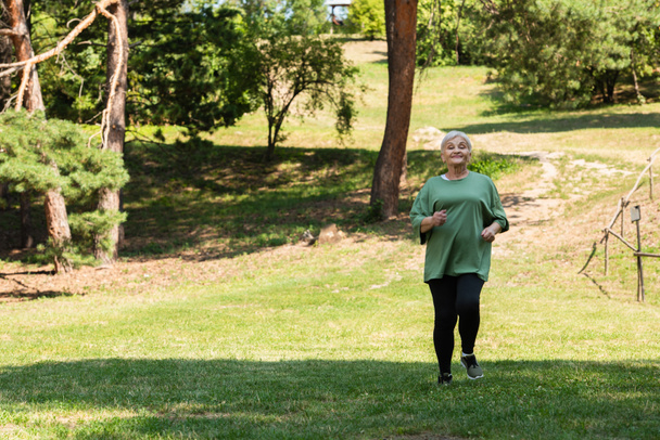 full length of senior woman with grey hair smiling and running on grass in park - Foto, Imagem