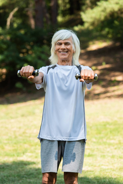 happy senior man in sportswear exercising with dumbbells in green park  - Foto, afbeelding