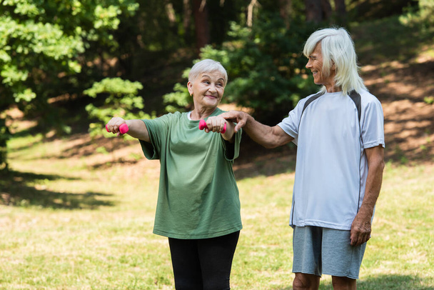 cheerful senior man in sportswear touching hand of wife working out with dumbbells in green park  - Photo, image