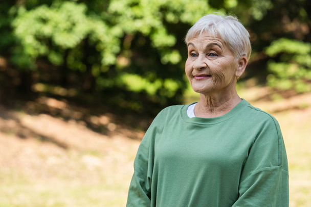 happy and retied woman with grey hair standing in green t-shirt in park - Fotografie, Obrázek