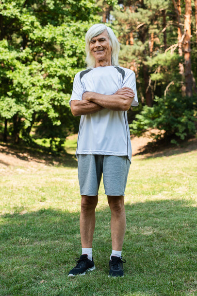 full length of happy senior man in sportswear standing with crossed arms in green park  - Foto, imagen