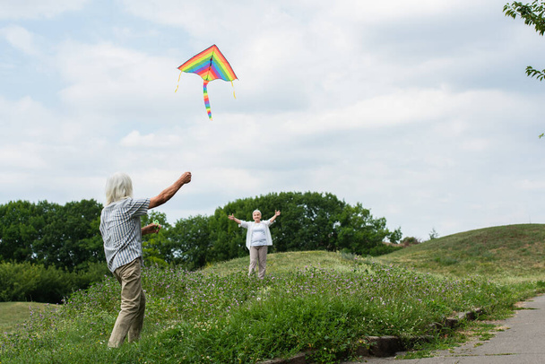 happy senior woman in casual clothes looking at husband playing with kite on green hill  - Foto, immagini