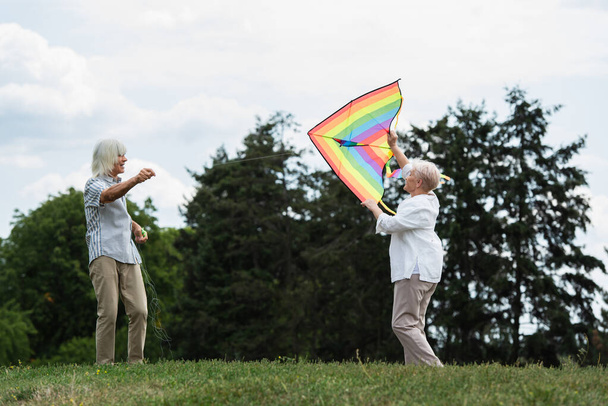 senior woman in casual clothes playing with kite near happy husband on green hill  - 写真・画像