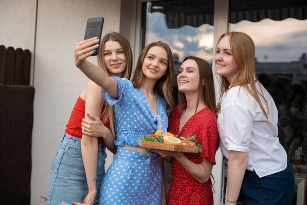 Group of young smiling good-looking women holding cut vegetables and fruits on wooden tray, stretching hand with smartphone, making selfie, enjoying. Friendship, celebration, hen-party, technology. - 写真・画像