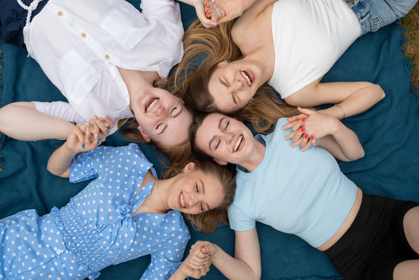 Top view of young laughing stunning women holding joining hands together, lying in circle on blue blanket with closed eyes, resting. Symbol of unity, support, collaboration, mutual help, relaxation. - Foto, imagen