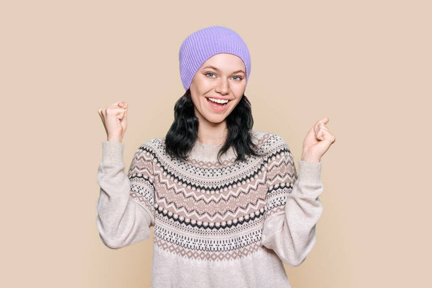 Portrait of happy young woman in sweater knitted hat celebrating winter holidays. Cheerful female showing joy victory gesture, yes sign, on brown color studio background. Winter Christmas season - Photo, Image