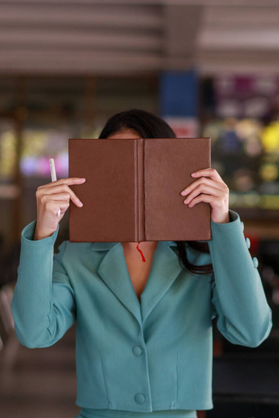 A woman wearing a green suit lifts a book to cover her face after reading for a while to block the light from entering her eyes because she wants to rest her eyes from reading for a long time. - Photo, image