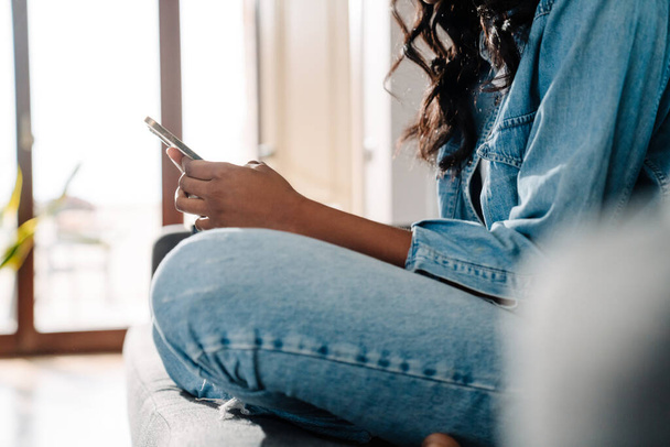 Black young woman using mobile phone while sitting on couch at home - Fotó, kép