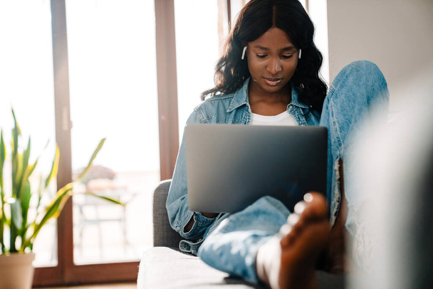 Black young woman in earphones using laptop while sitting on couch at home - Fotó, kép