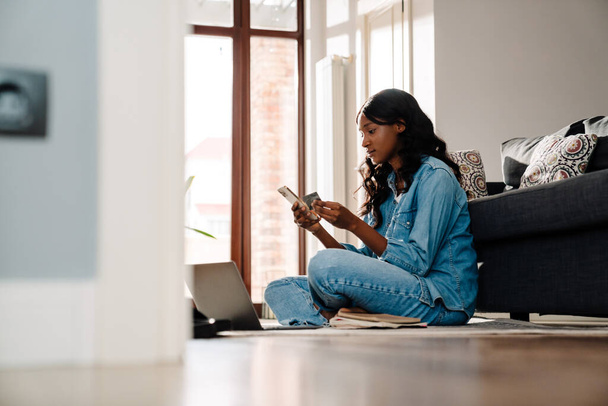 Black young woman using cellphone and credit card while sitting on floor at home - Zdjęcie, obraz