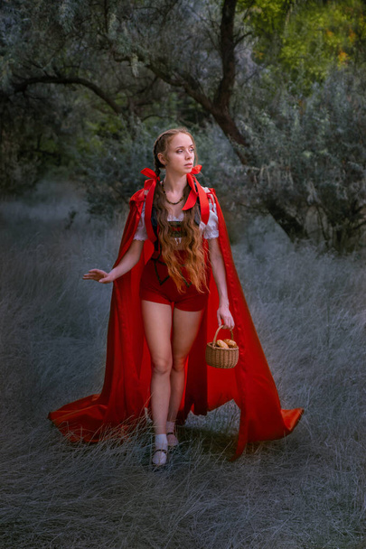 милый фантастический образ сказочного персонажа, таинственная рыжеволосая девушка с длинным трепещущим алым ярко-красным плащом стоит в лесу, Портрет героини детской книги в лесу - Фото, изображение