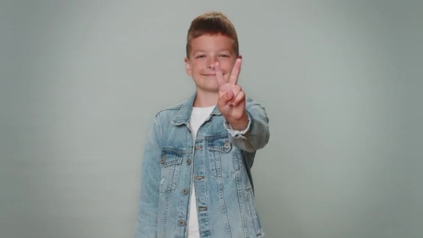 Toddler boy showing victory sign, hoping for success and win, doing peace gesture, smiling with kind optimistic expression. Young teenager children child kid indoor isolated on gray studio background - Felvétel, videó