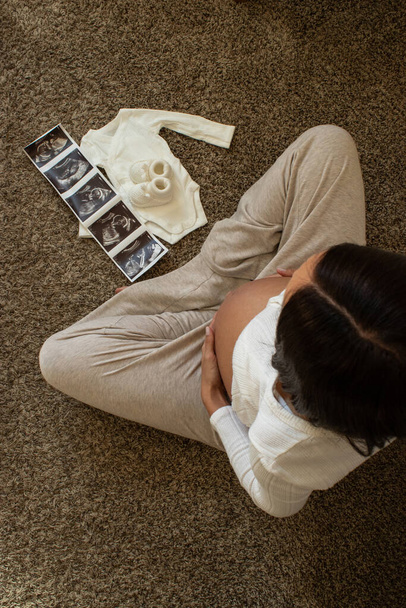 young woman sitting on the floor with ultrasound pictures and baby cloths, holding belly - 写真・画像