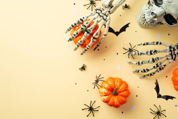 Halloween decor concept. Top view photo of scary skull skeleton hands holding pumpkins spiders bat silhouettes and confetti on isolated beige background with copyspace - 写真・画像