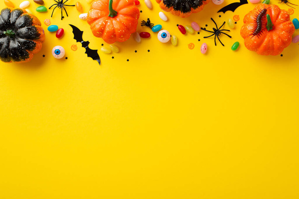 Halloween decorations concept. Top view photo of pumpkins spooky eyeballs centipede color candies spiders and confetti on isolated yellow background with empty space - 写真・画像