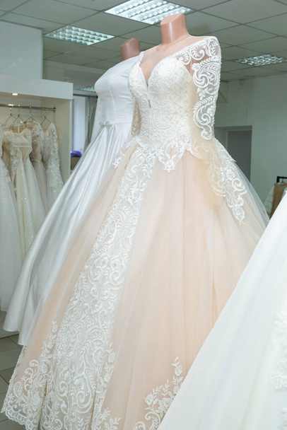 A beautiful cream wedding dress on a mannequin. A wedding dress on the background of other wedding dresses in a wedding salon. - Foto, Imagen