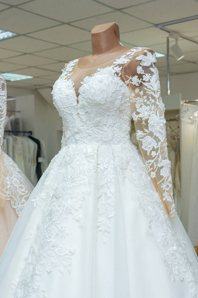 A beautiful white wedding dress on a mannequin. A close-up of a dress against other wedding dresses in a bridal shop. - Valokuva, kuva
