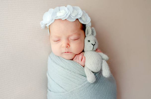 Newborn baby swaddled in fabric holding knitted bunny toy and sleeping portrait. Infant child kid napping studio photoshoot - Zdjęcie, obraz