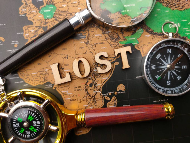 A flat lay with a magnifying glass, compass and a wooden word LOST on a world map background - Фото, зображення