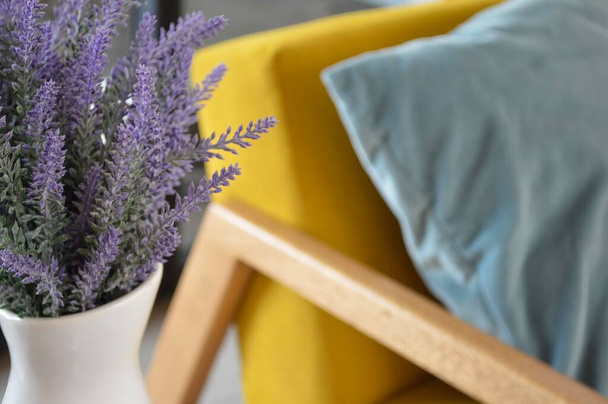 A closeup of a bunch of lavender in a vase by a yellow chair. - 写真・画像