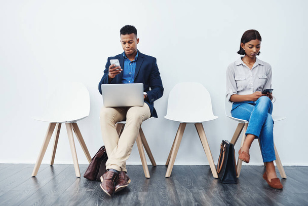 Were starting to get a bit nervous now. Full length shot of two young businesspeople sitting down on chairs and using their tech devices against a grey background - Fotoğraf, Görsel