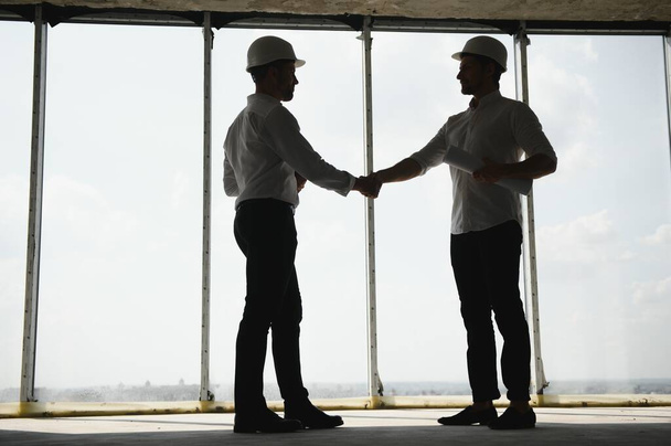 A front view of two smart architects with white helmets reviewing blueprints at a construction site on a bright sunny day. - Φωτογραφία, εικόνα