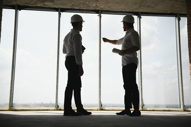 A front view of two smart architects with white helmets reviewing blueprints at a construction site on a bright sunny day. - Photo, Image