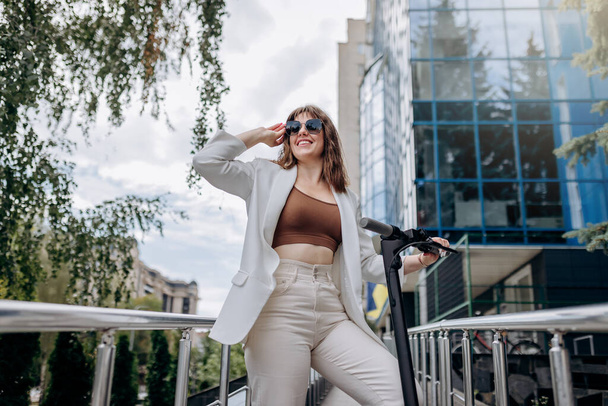 Красива молода жінка в сонцезахисних окулярах і білому костюмі стоїть зі своїм електричним скутером біля сучасної будівлі і дивиться в сторону
  - Фото, зображення