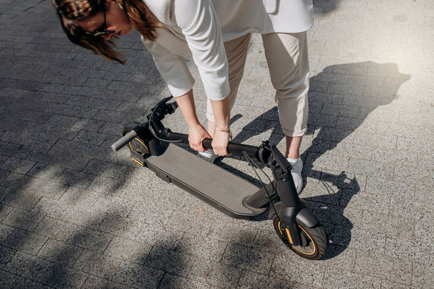 Close up of woman in white suit folding her electro scooter after ride while standing in city parkland - Valokuva, kuva