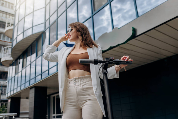 Красивая молодая женщина в солнечных очках и белом костюме, стоящая со своим электрическим скутером рядом с современным зданием и глядя в сторону  - Фото, изображение