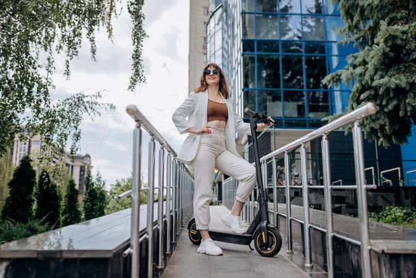 Красива молода жінка в сонцезахисних окулярах і білому костюмі стоїть зі своїм електричним скутером біля сучасної будівлі і дивиться в сторону
  - Фото, зображення