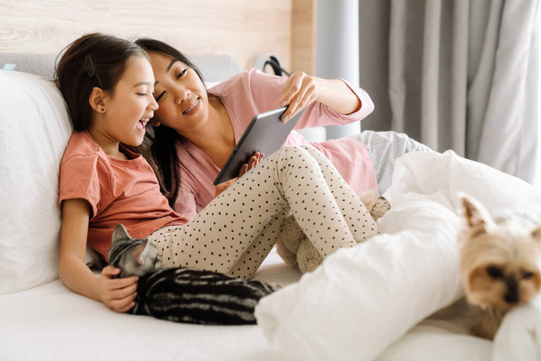 Asian mother and daughter using tablet computer while sitting on bed at home - Fotografie, Obrázek
