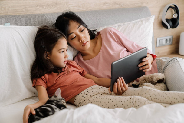 Asian mother and daughter using tablet computer while sitting on bed at home - Fotoğraf, Görsel
