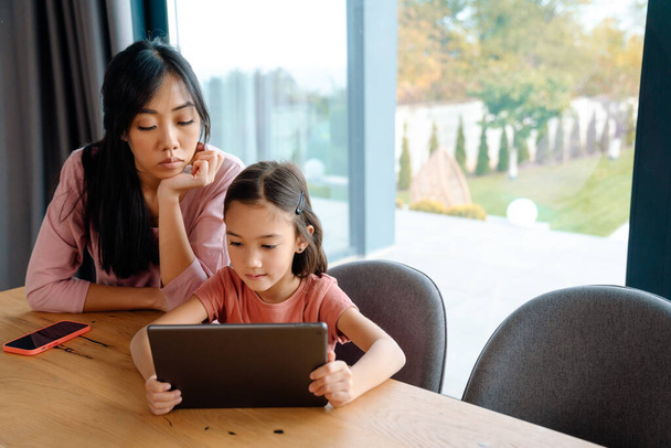 Asian woman and her daughter using tablet computer while sitting at table in home - Foto, afbeelding