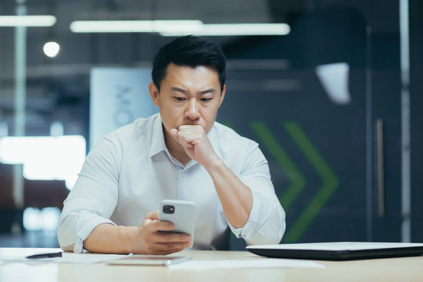 Confused Asian guy holding smart phone feels concerned thinking over received message. Mobile phone everyday usage, unpleasant news, waiting for important call, low signal device problems concept - Φωτογραφία, εικόνα