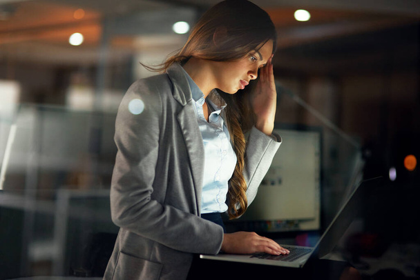 Stressed, worried and tired female entrepreneur typing on a laptop and working overtime late at night. Frustrated and overworked young businesswoman with a headache doing a project with a deadline. - 写真・画像