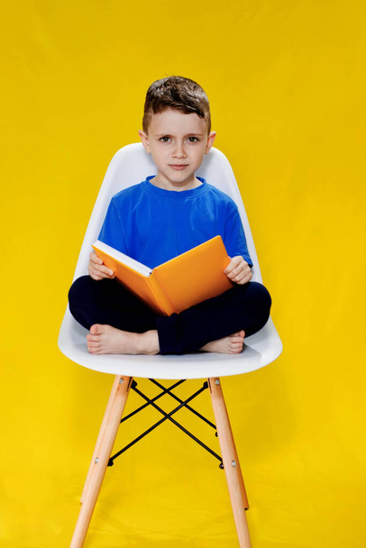 Little cheerful blond green-eyed boy 5-6 years old in a stylish blue T-shirt holding book and reading on yellow wall background, children's studio portrait. - Foto, imagen
