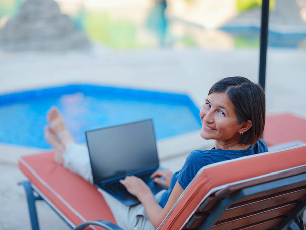 Young business woman working at computer by hotel pool. Young lady downshifter working at laptop and enjoys and relaxed environment, working day. Online freelance work on vacation. - Photo, image