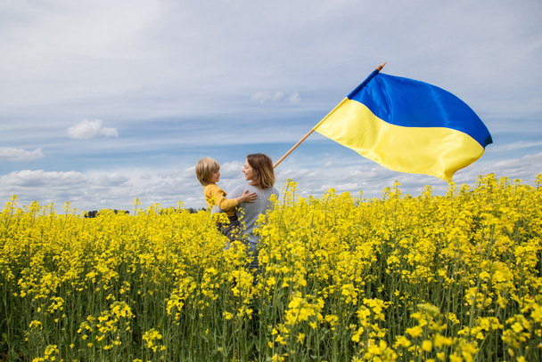 mother with son in her arms, with large satin flag of Ukraine, stand among blooming yellow rapeseed field. Glory to Ukraine. Education of patriotism. Pride to be Ukrainian, national independence - Fotografie, Obrázek