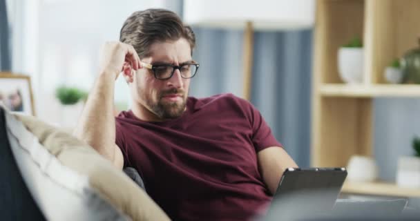 Stressed man with a bad headache while on tablet reading, watching or sitting on a sofa at home. Unhappy male suffering from a painful migraine, feeling mentally drained, eyes strained or tired. - Filmmaterial, Video