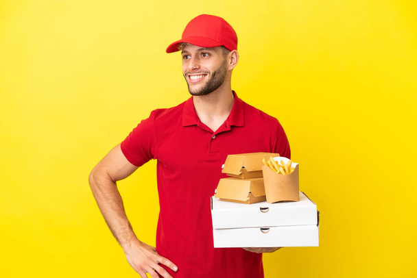 pizza delivery man picking up pizza boxes and burgers over isolated background posing with arms at hip and smiling - Foto, Bild