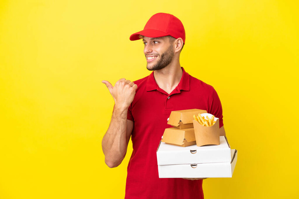 Pizzalieferant nimmt Pizzakartons und Burger über isoliertem Hintergrund entgegen und zeigt auf die Seite, um ein Produkt zu präsentieren - Foto, Bild