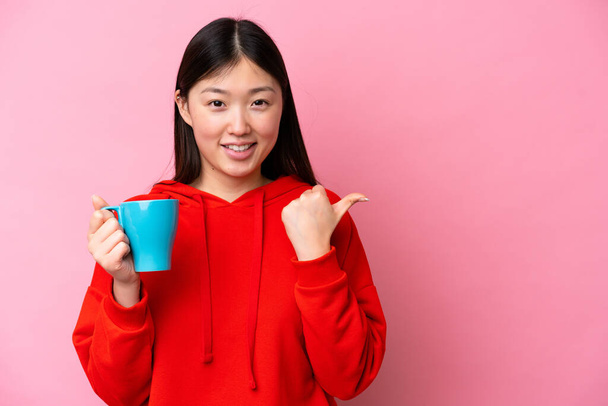 Молодая китаянка держит чашку кофе изолированы на розовом фоне указывая в сторону представить продукт - Фото, изображение