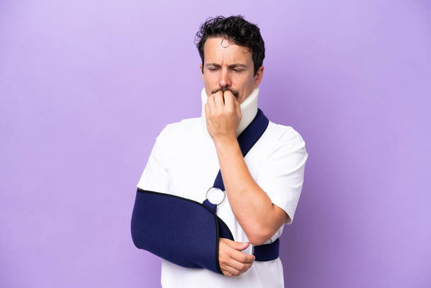 Young caucasian man wearing a sling and neck brace isolated on purple background having doubts - Photo, image