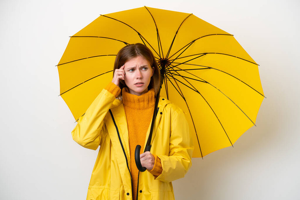 Молода англійська жінка з дощовитим пальто і парасолька ізольовані на білому фоні мають сумніви і мислення - Фото, зображення