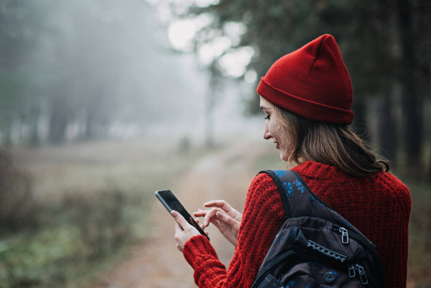 Travel Apps. Young woman traveler with backpack holding smartphone device in hands while walking in pine forest. Happy woman enjoying new adventure and using travel apps or gps navigation in cell - 写真・画像