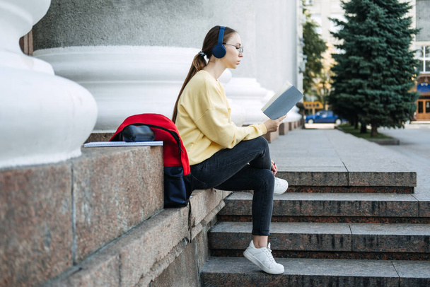 Εξωτερική Πορτρέτο του κοριτσιού ευτυχής φοιτητής, νεαρή γυναίκα ακούγοντας μαθήματα ήχου, μαθήματα, podcast, μουσική μέσω ακουστικών με βιβλίο, σημειωματάριο στο δρόμο. - Φωτογραφία, εικόνα