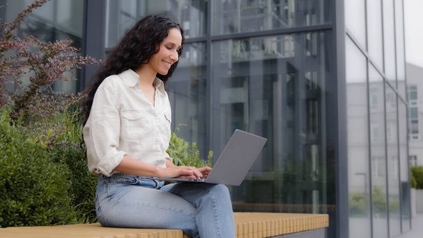 Улыбающаяся деловая женщина сидит на скамейке рядом с офисным зданием на террасе с ноутбуком, общаясь с клиентами, пишет статья. Электронное обучение для девочек с использованием беспроводного компьютера с бесплатным Wi-Fi - Фото, изображение
