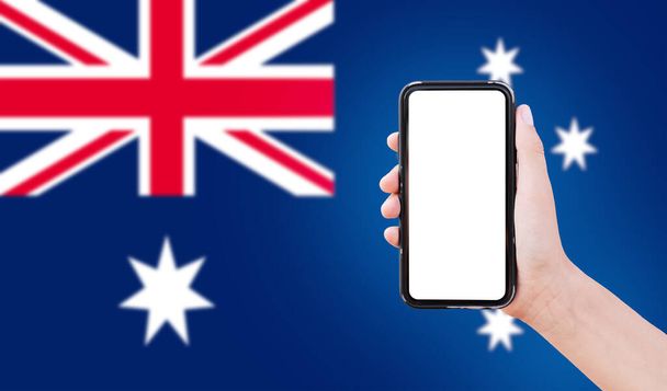 Close-up of male hand holding smartphone with blank on screen, on background of blurred flag of Australia. - Fotó, kép